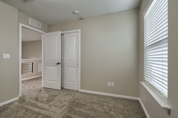 A small empty bedroom with carpet, an open white door, and a window with blinds